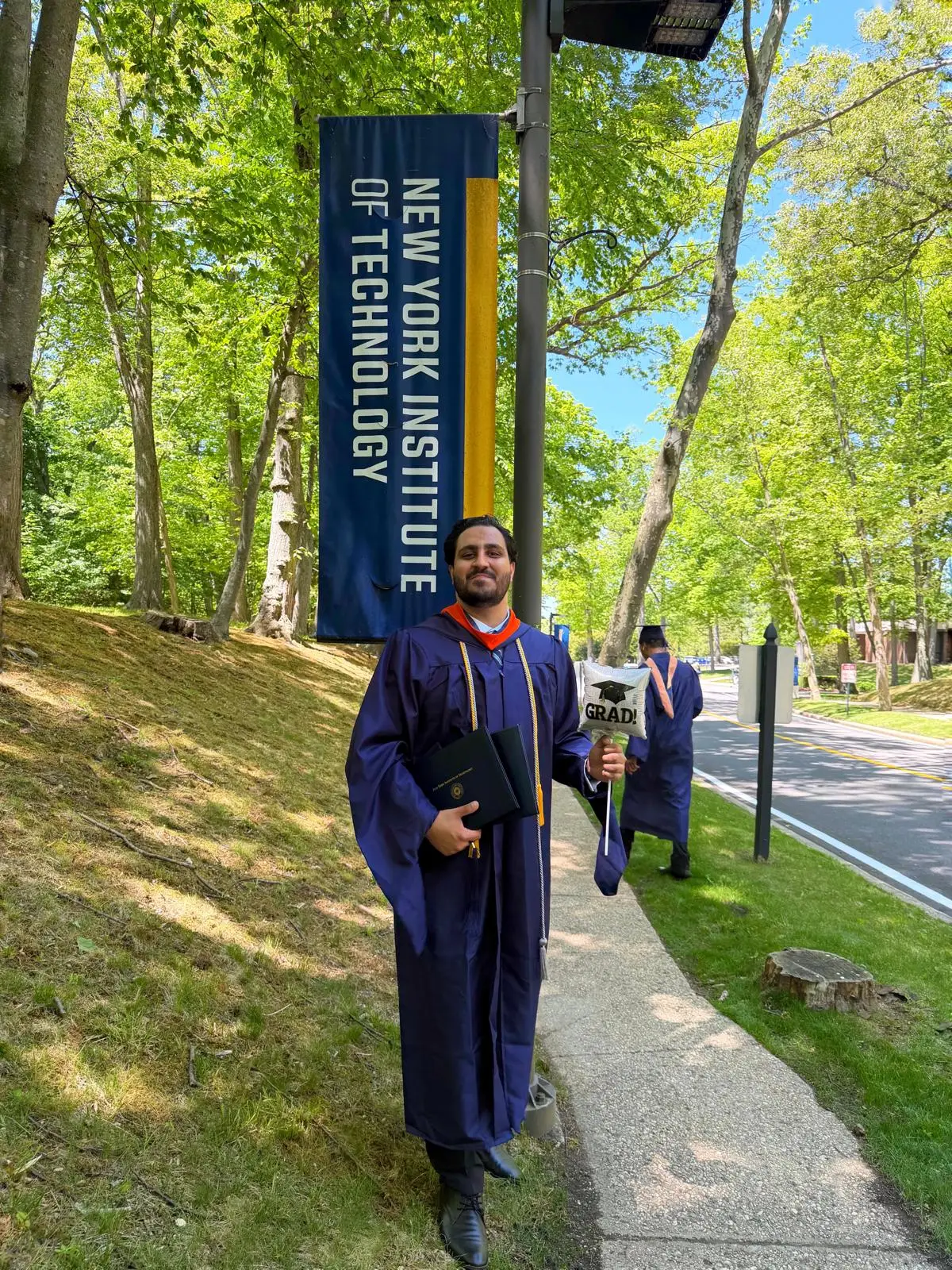 Zulqarnain in regalia beside the New York Institute of Technology banner at commencement